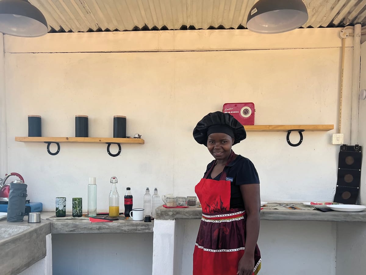 Izzie, Wild Bhiza's chef, wearing traditional attire in rustic outdoor kitchen preparing authentic African refreshments for horse riding tour guests near Victoria Falls