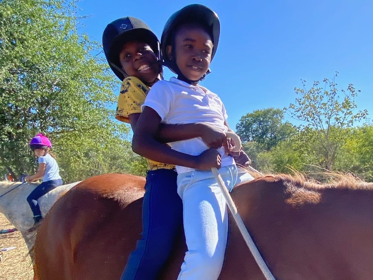 Two smiling local Zimbabwean children riding together on horseback during Wild Bhiza youth education program in Chisuma Village near Victoria Falls