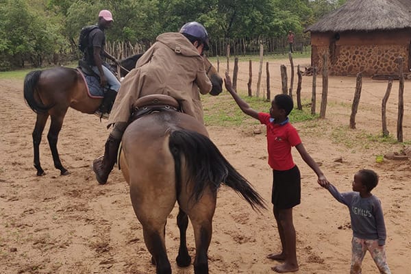 horse-riding-village-tour-local-children-victoria-falls-zimbabwe.jpg Tourists on horseback greeting local African children in traditional village near Victoria Falls Zimbabwe showcasing authentic cultural horse riding experience